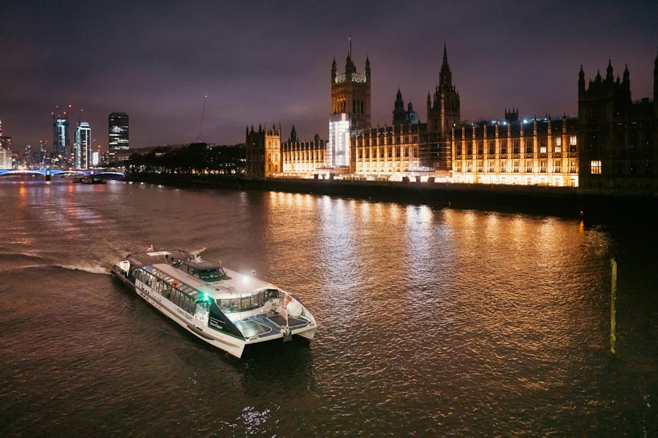 A boat sails on a river at night with the illuminated British Parliament building in the background.
