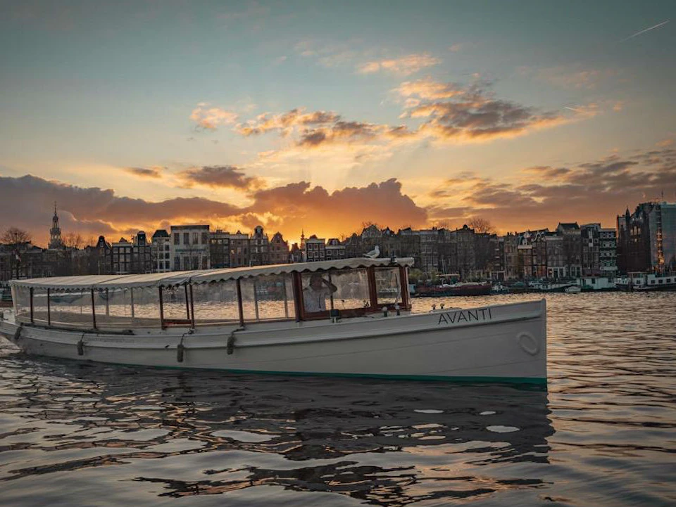 A white boat named "Avanti" floats on water at sunset with a cityscape in the background under a partly cloudy sky.