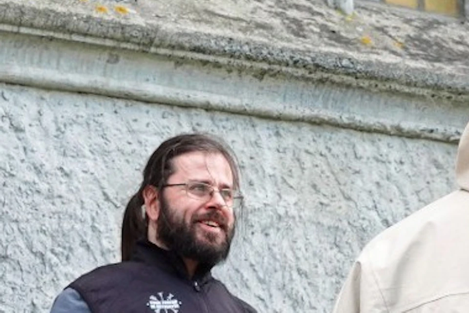 Three people stand near a stone building with a stained glass window, engaged in conversation.
