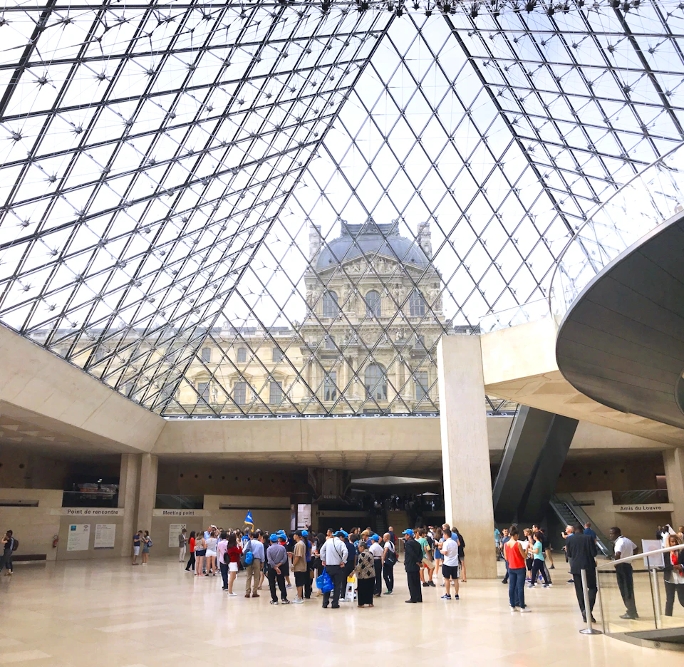 A group of people stand under the glass pyramid entrance of the Louvre Museum, with the historic building in the background.