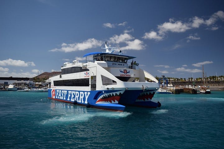 Velký modro-bílý trajekt s nápisem „Fast Ferry“ na boku, blížící se k doku, pod částečně zataženou oblohou.