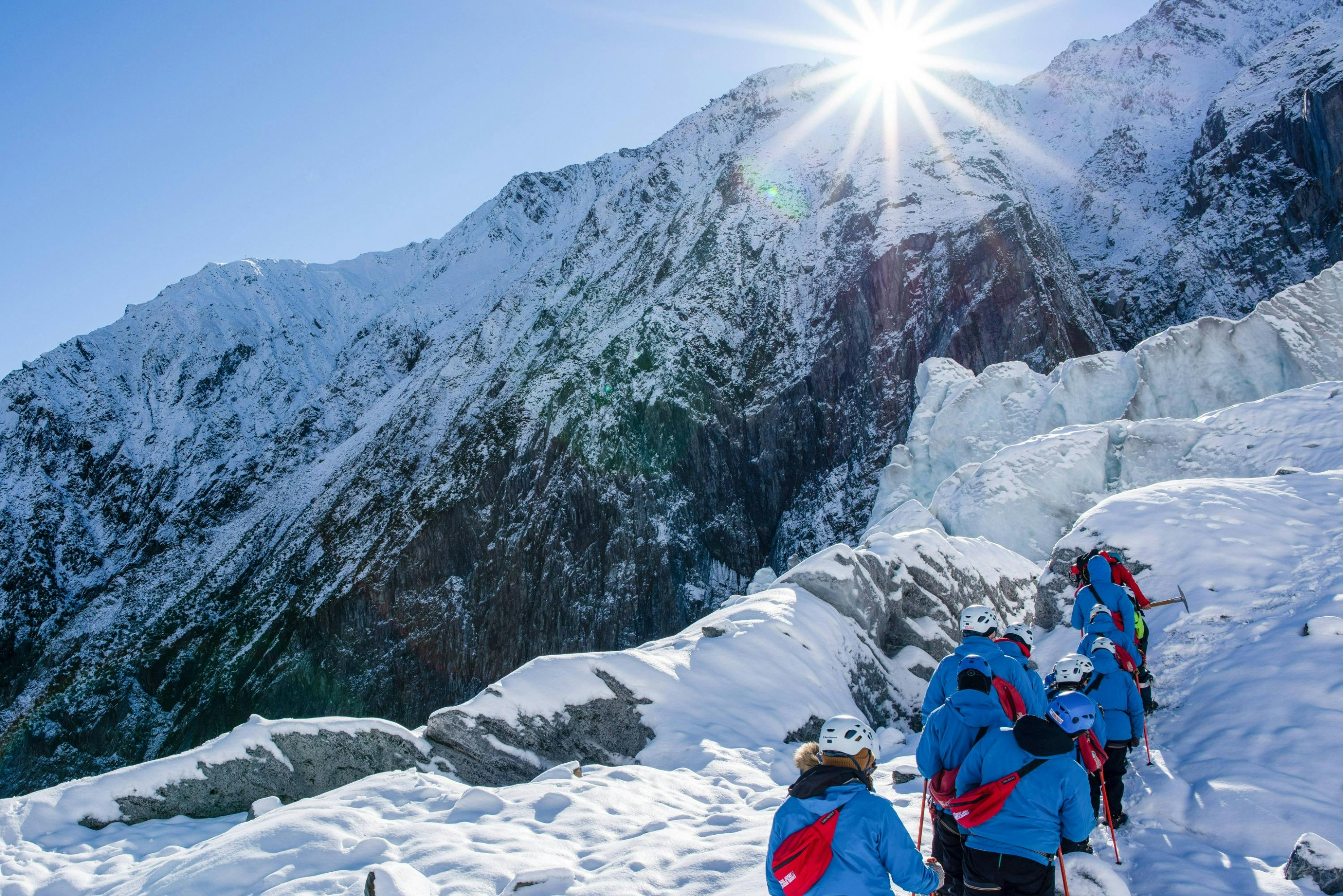 A group of people in blue jackets and helmets hiking on a snowy mountain under bright sunlight.