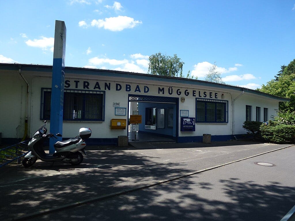 Entrance to "Strandbad Müggelsee" with a scooter parked nearby, located by the street on a sunny day.