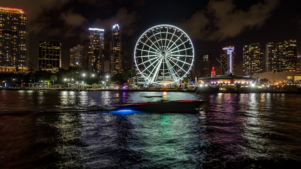 Illuminated Ferris wheel by a waterfront at night, with city skyscrapers and a passing boat reflecting lights on the water.