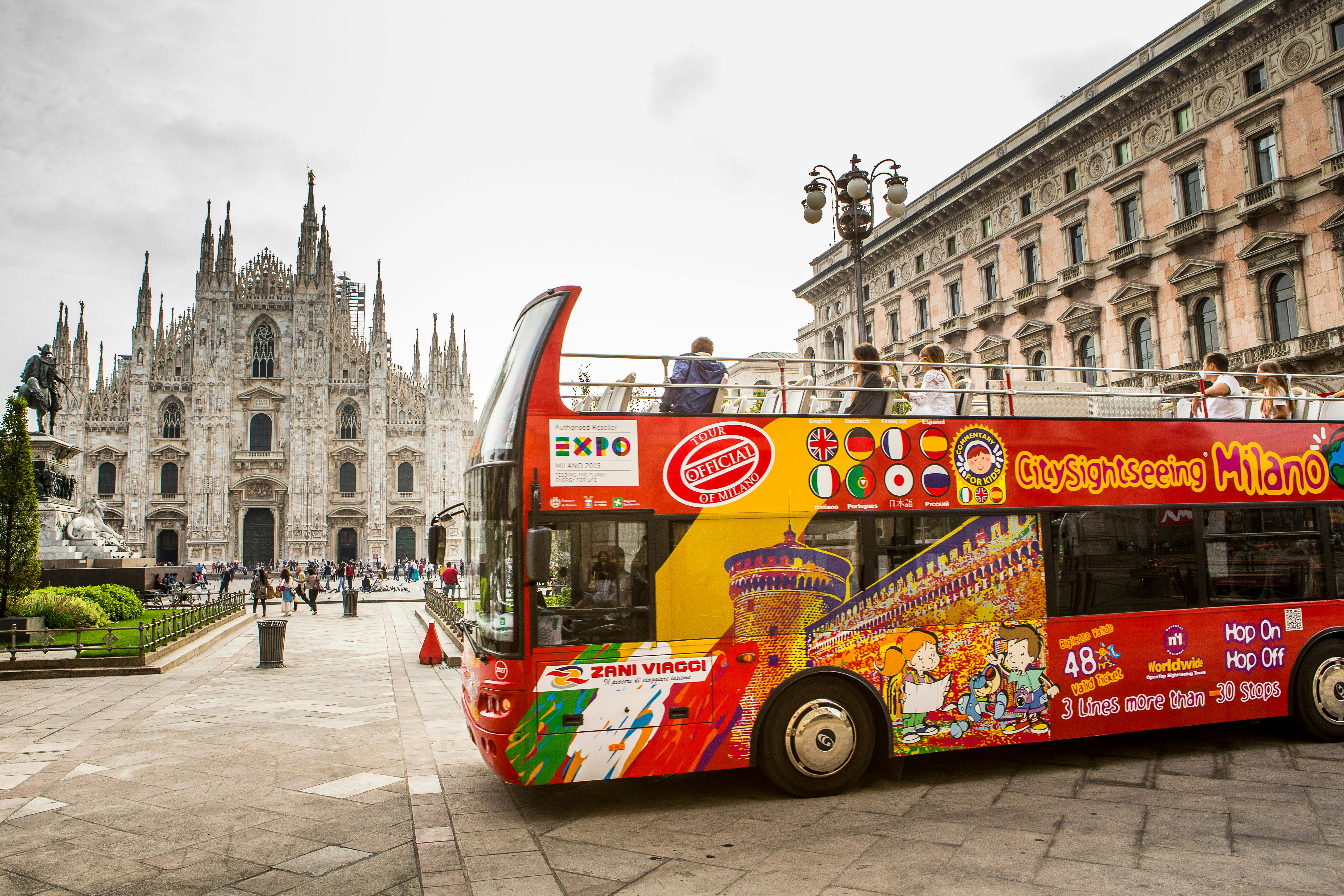 Un colorato autobus turistico a due piani a Milano con persone sul ponte superiore, il Duomo di Milano e una statua visibile sullo sfondo.