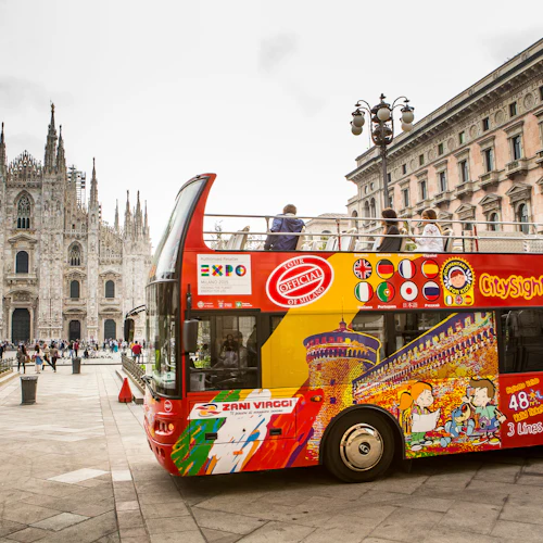 En färgglad dubbeldäckad sightseeingbuss i Milano med människor på övre däck, Milanos katedral och en staty synlig i bakgrunden.
