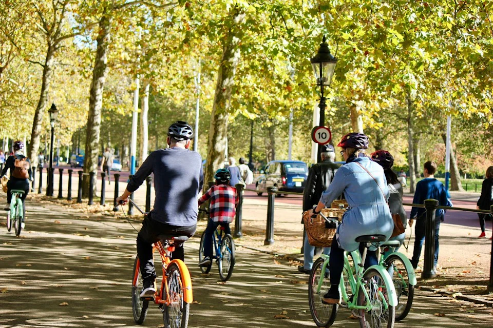 People ride bicycles on a tree-lined path in a park. Light poles and a 10 mph speed sign are visible in the background.