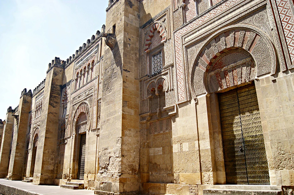 Exterior of a historic building with intricate arches, geometric patterns, and weathered stone walls under a clear sky.