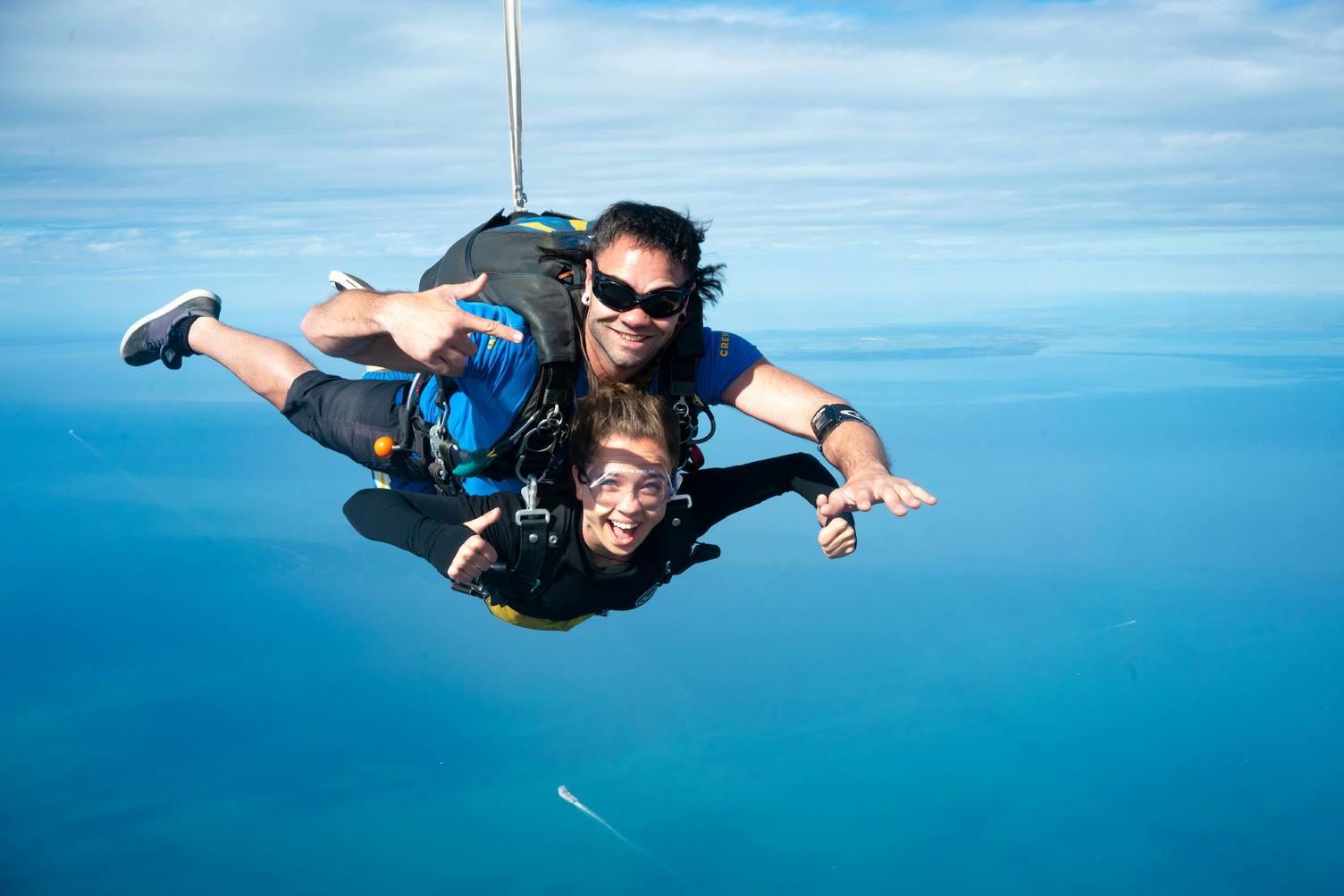 Δύο άνθρωποι κάνουν ελεύθερη πτώση με αλεξίπτωτο, φορώντας ιμάντες και γυαλιά, με φόντο τον γαλάζιο ουρανό και τον ωκεανό.