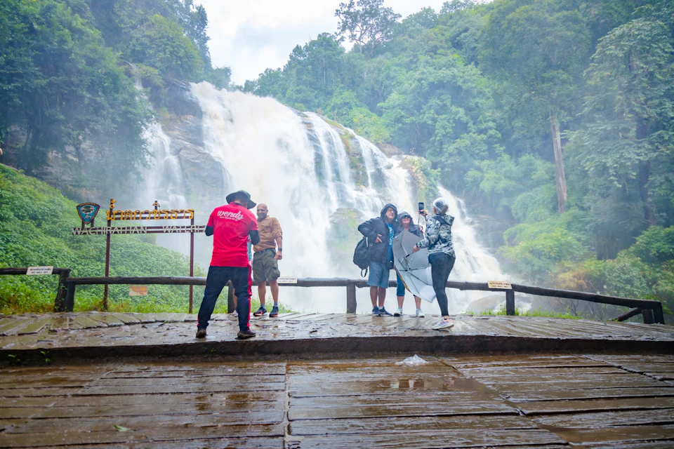 People pose for photos in front of a large waterfall surrounded by lush greenery, on a wet, wooden viewing platform.