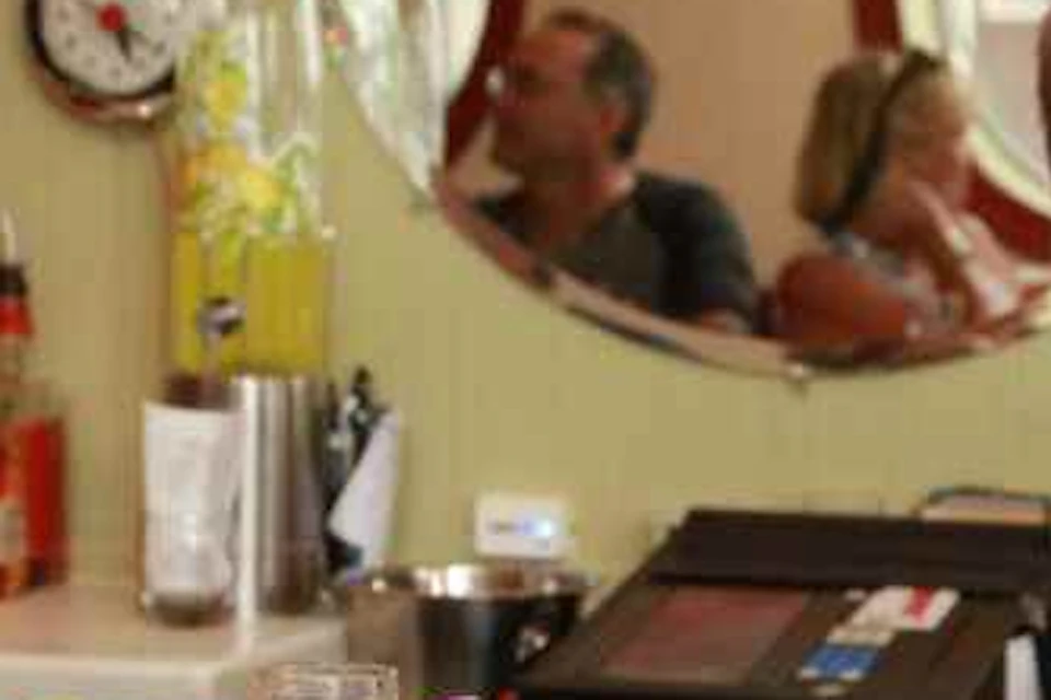 A smiling man at a bar holding a cocktail, with bottles and a small mirror behind him reflecting other patrons.