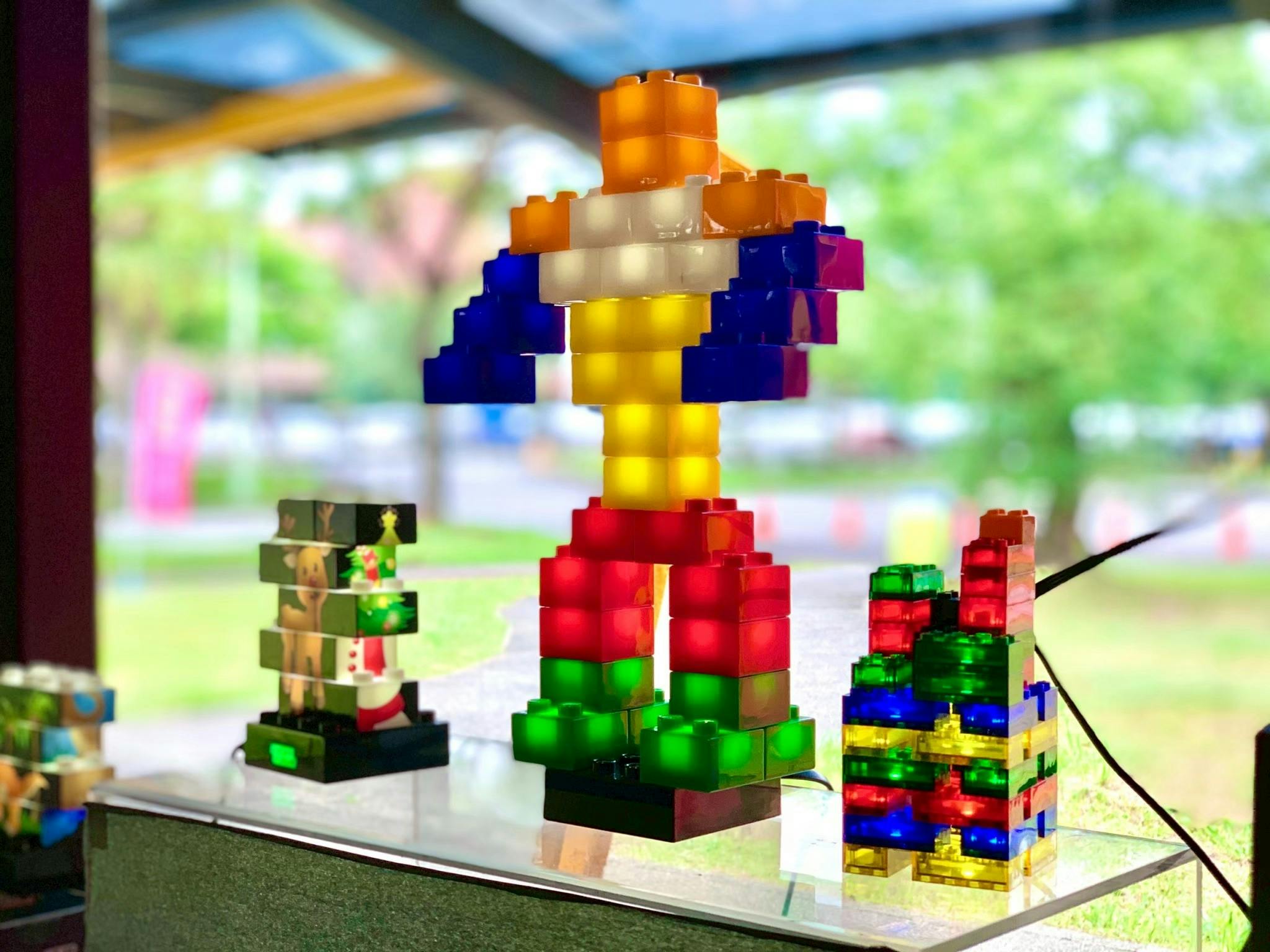 Colorful constructions made of translucent building blocks are displayed on a table, with an outdoor setting in the background.