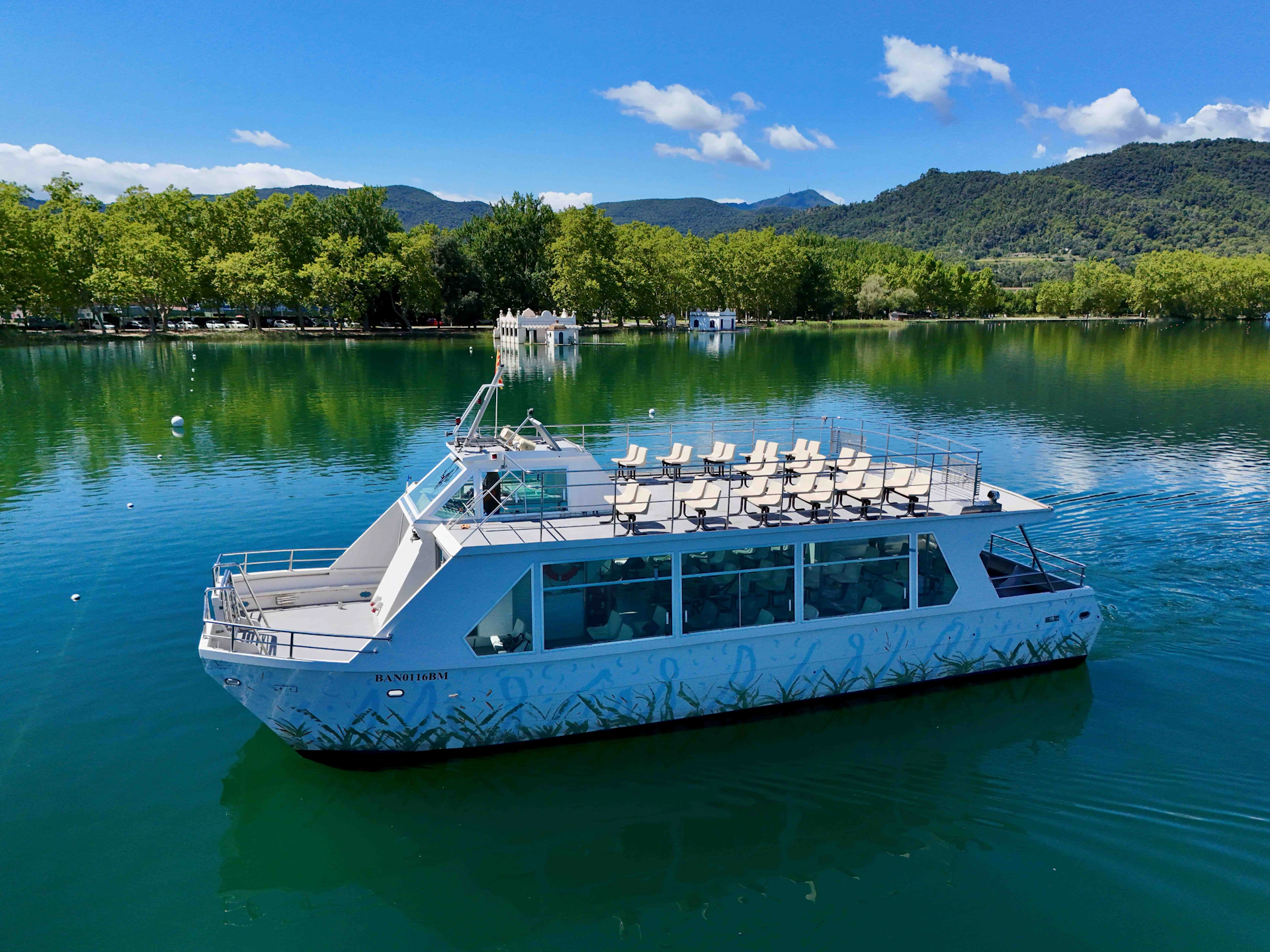 Modern white boat with upper-deck seating, floating on a calm lake surrounded by lush trees and distant mountains.