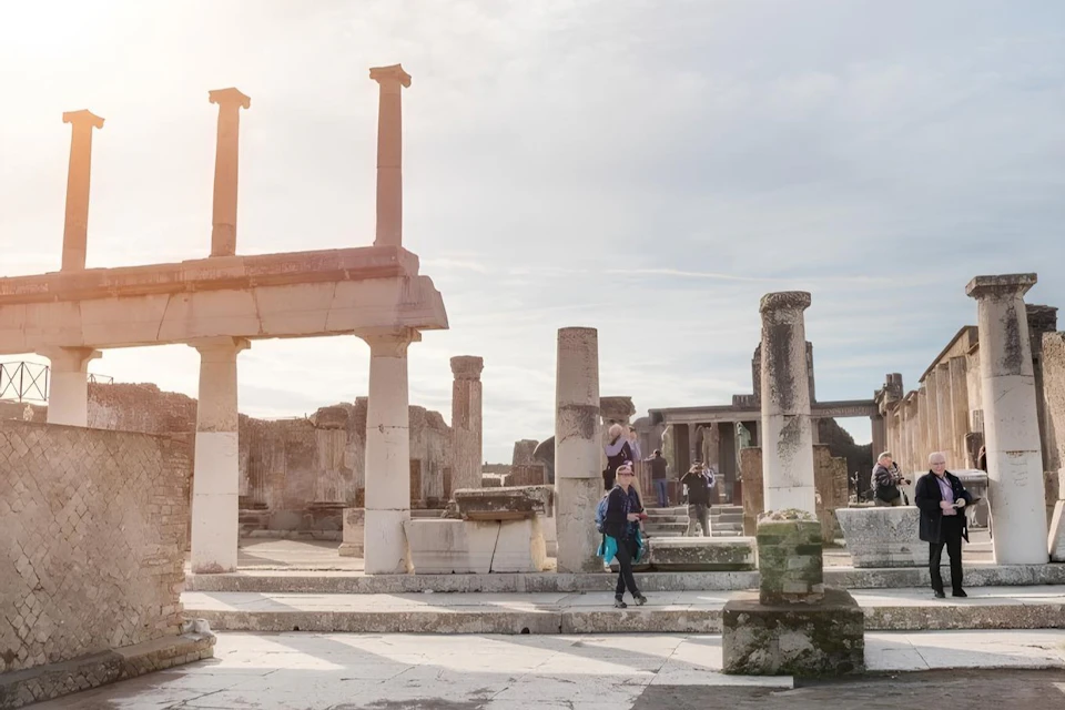 People walking among ancient ruins with stone columns and structures under a cloudy sky.