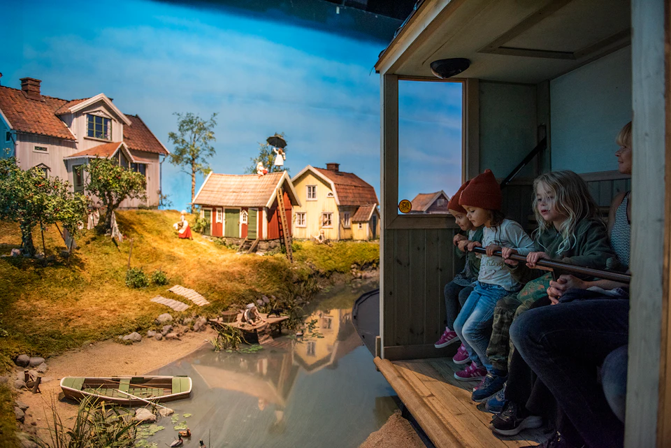 Four children and an adult seated in a wooden boat, looking at miniature houses and scenery through an open window.