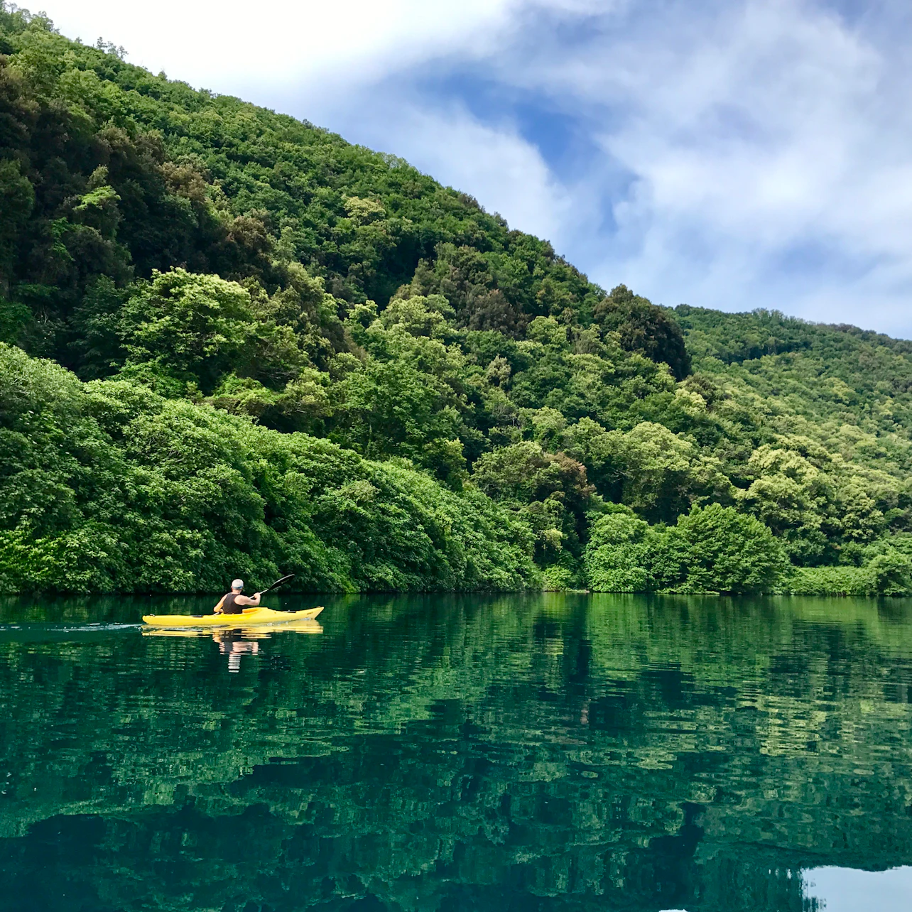 A person kayaks on a calm turquoise lake surrounded by dense green forested hills under a partly cloudy sky.