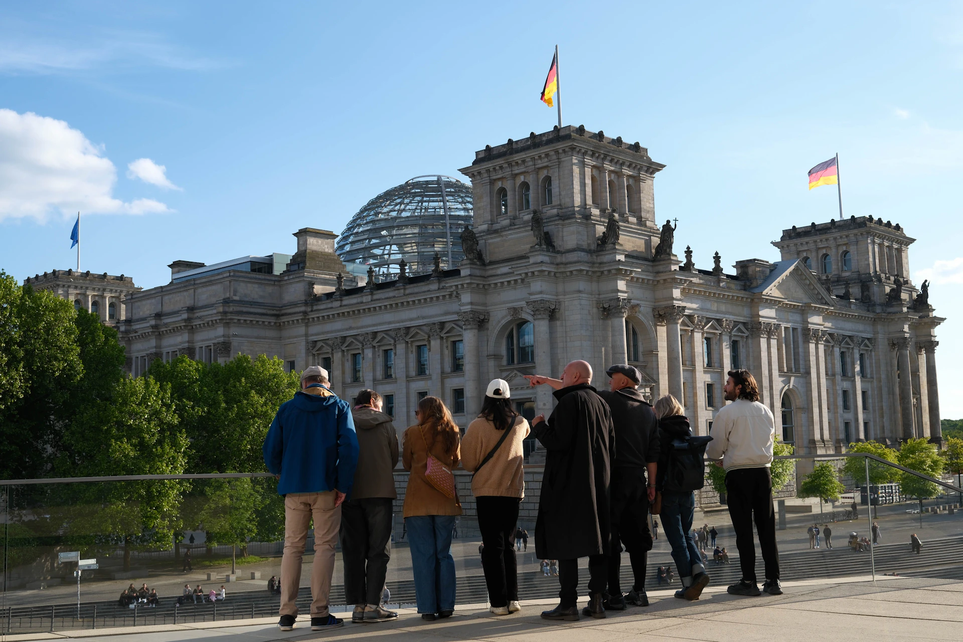 A group of people standing outdoors, facing the Reichstag building with a glass dome and a German flag on top.