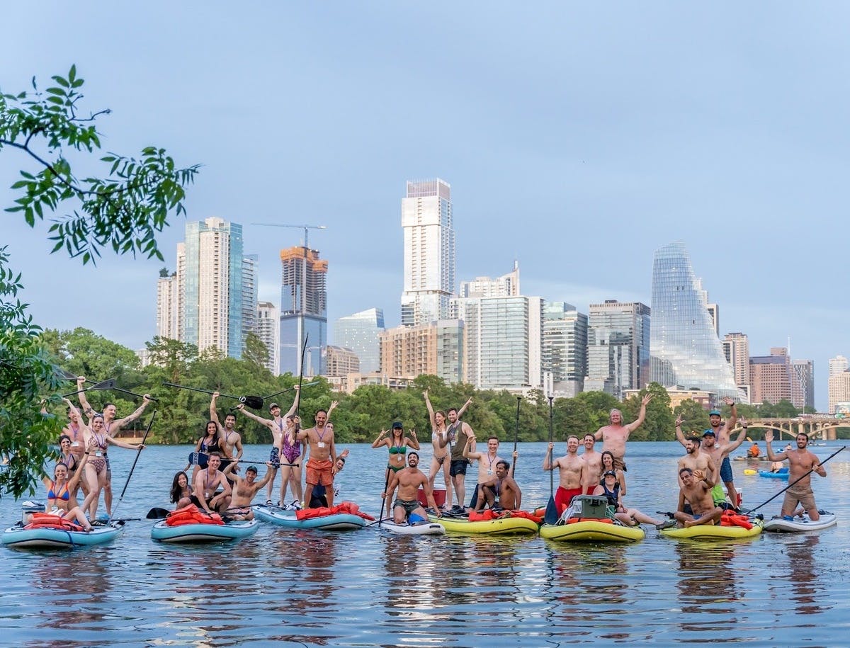 A group of people on paddleboards in a lake, with a city skyline and greenery in the background.