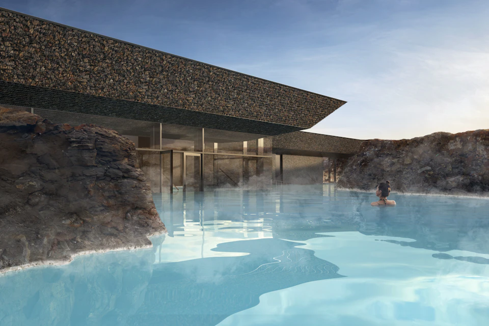 A modern building with stone walls overlooks a blue geothermal pool surrounded by rocky formations; a person is swimming.