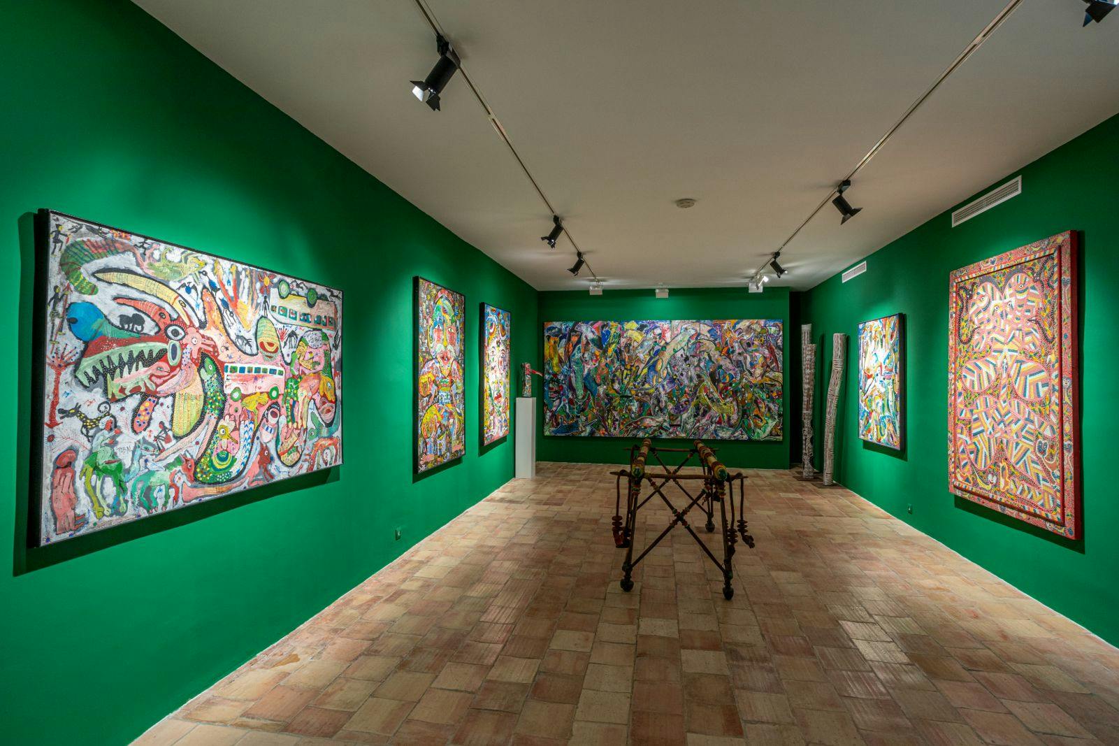 Art gallery with green walls displaying colorful abstract paintings and sculptures. Spotlights illuminate the artworks.