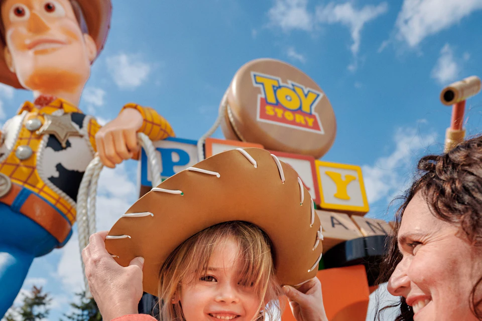 A child wearing a cowboy hat, being helped by an adult, standing in front of a large statue of Woody from \*Toy Story\* in an outdoor park.