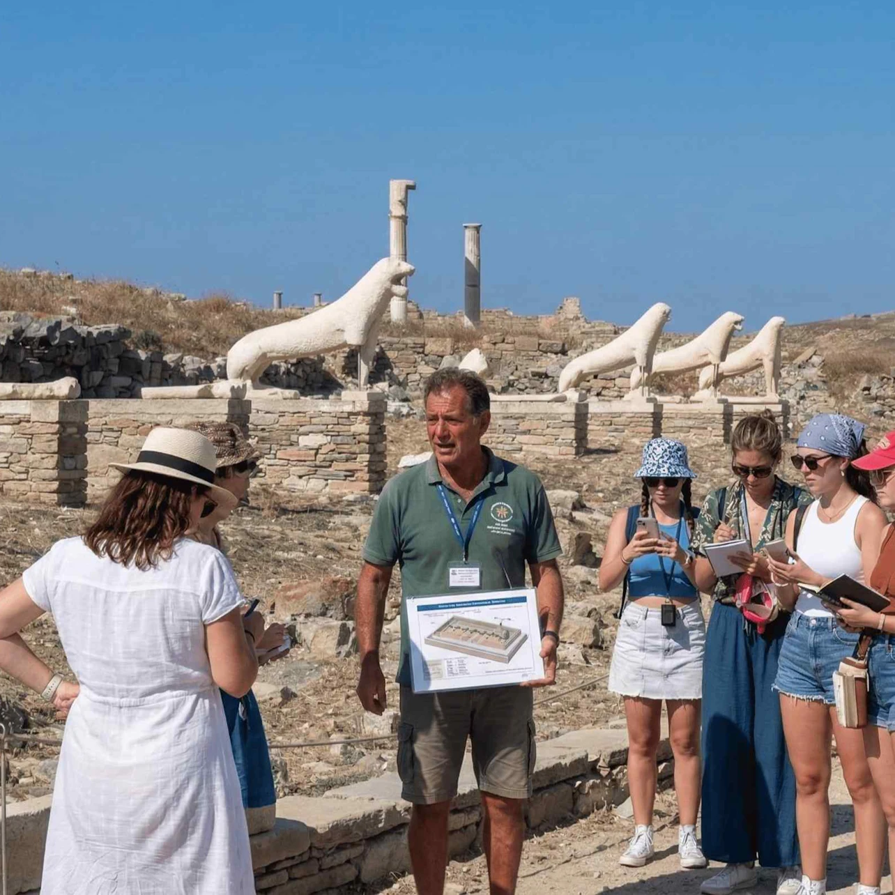 A guide showing a map to a group of tourists next to ancient sculptures of lions under a clear blue sky.