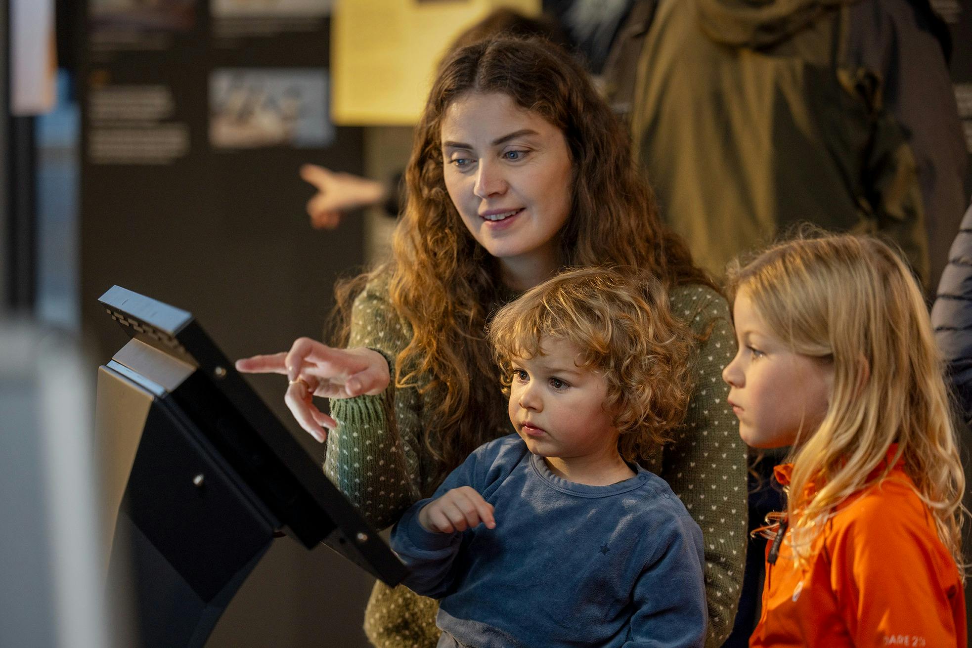 En kvinde med krøllet hår sidder med to børn, den ene i en orange jakke og den anden med krøllet hår, og kigger på en tablet.