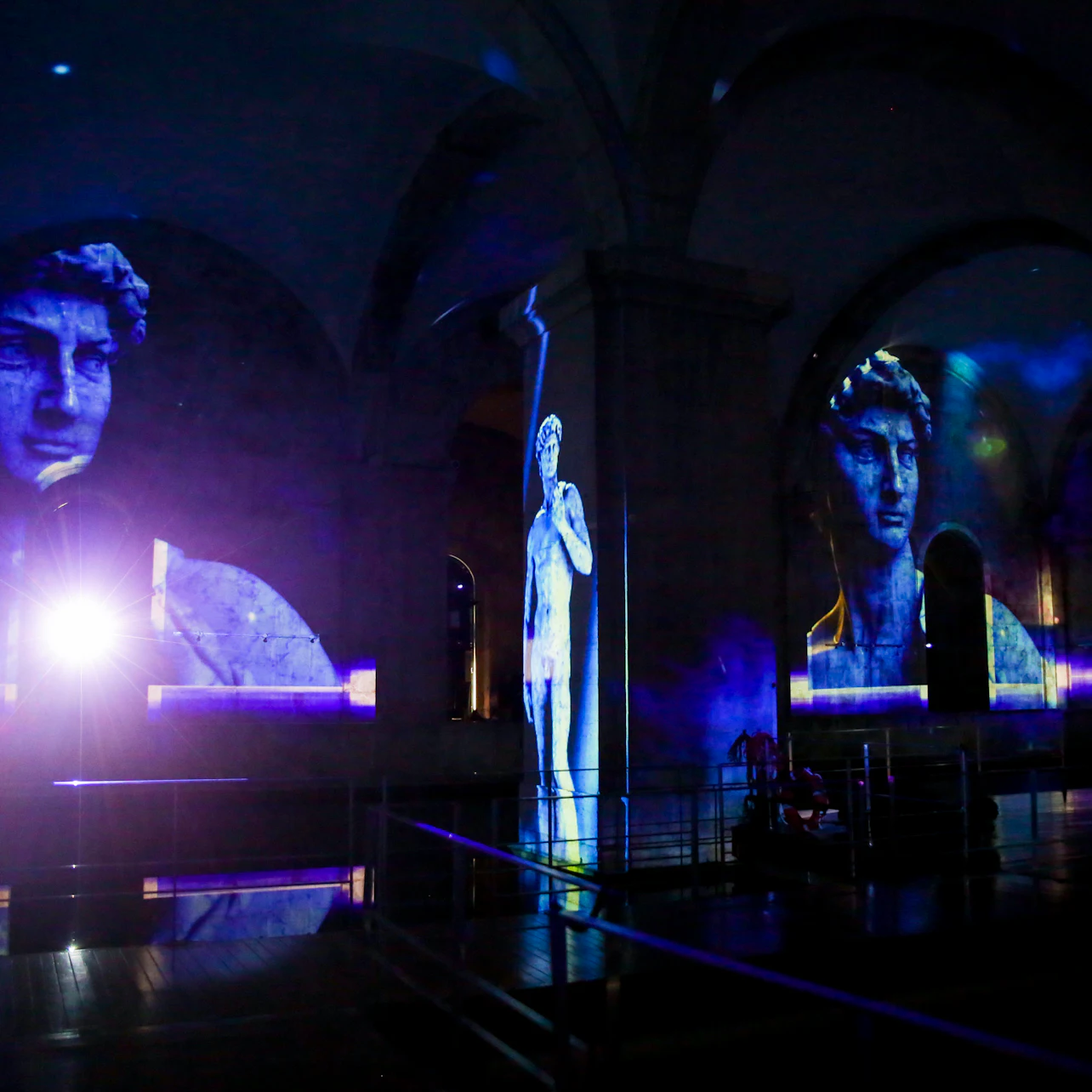 A museum exhibit displaying projected images of Michelangelo's David on arched walls, with a spotlight illuminating the scene.