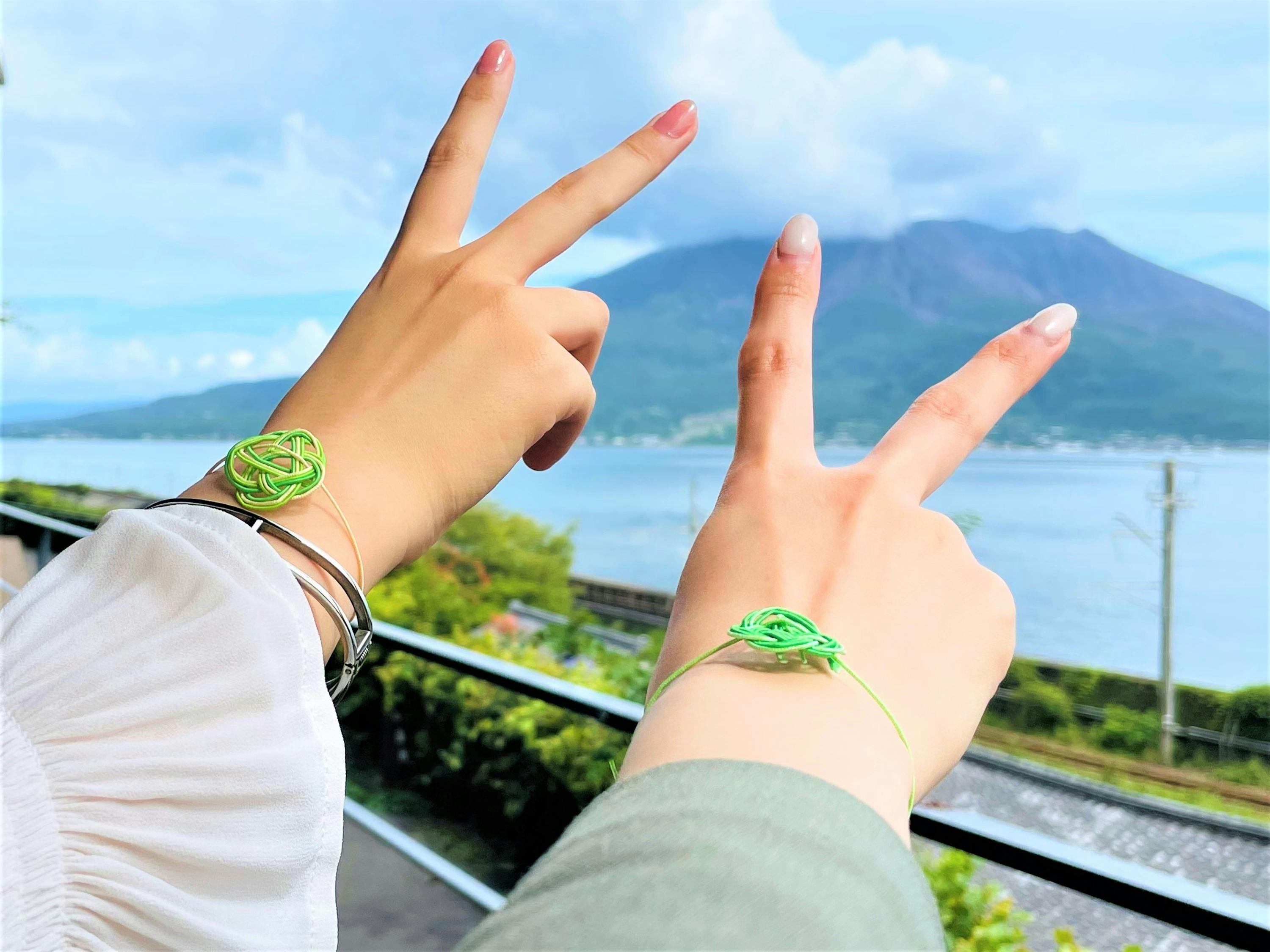 Two hands making peace signs, each wearing a green decorative bracelet, with a scenic mountain and lake background.