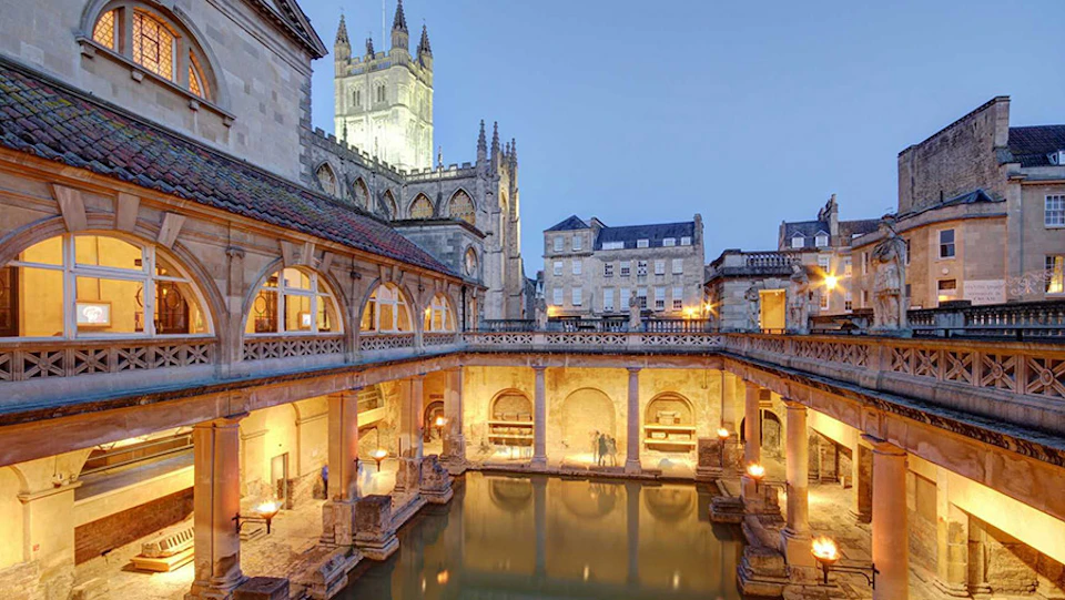 Historic Roman baths with a central pool surrounded by arched structures, with a tall church tower visible in the background.