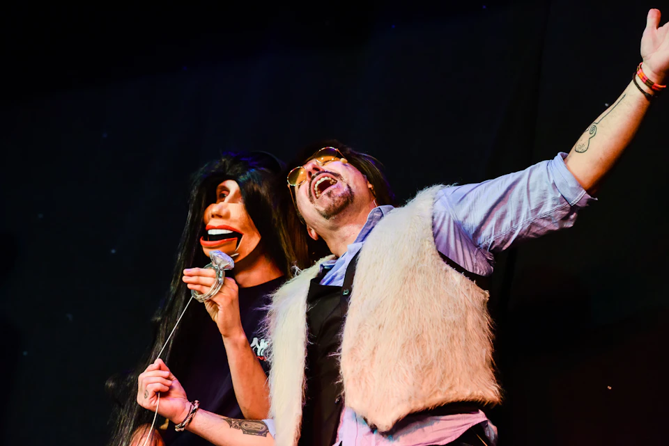 A person wearing a fur vest and sunglasses performs with a dark-haired ventriloquist dummy on stage.