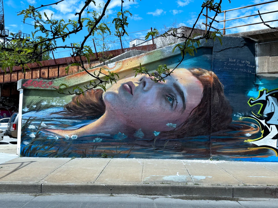 A street mural depicts a realistic painting of a woman's face partially submerged in water, with plants and sky above.