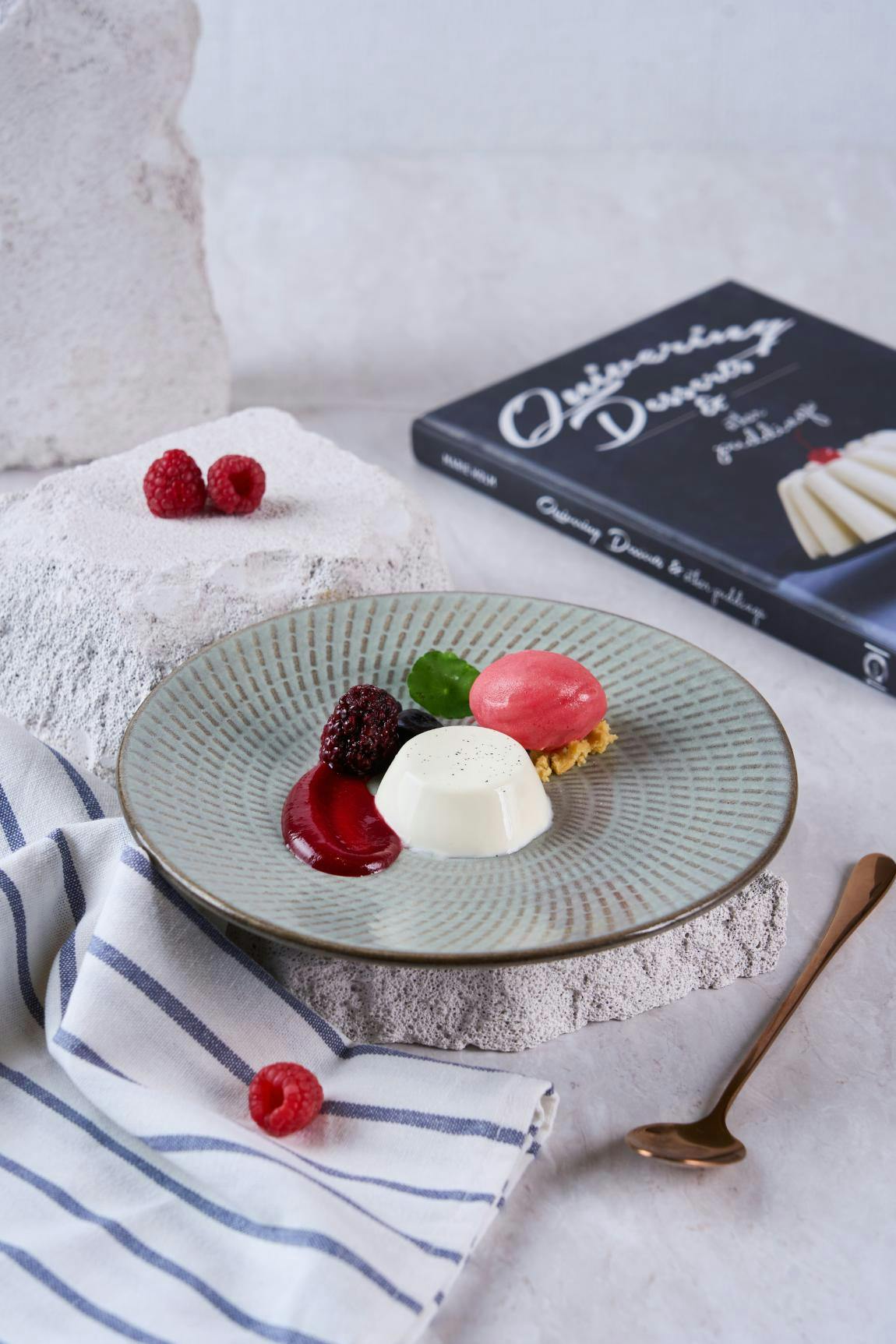 A plate with panna cotta, fruit garnishes, and sauce, placed on a textured surface with a cookbook and napkin nearby.