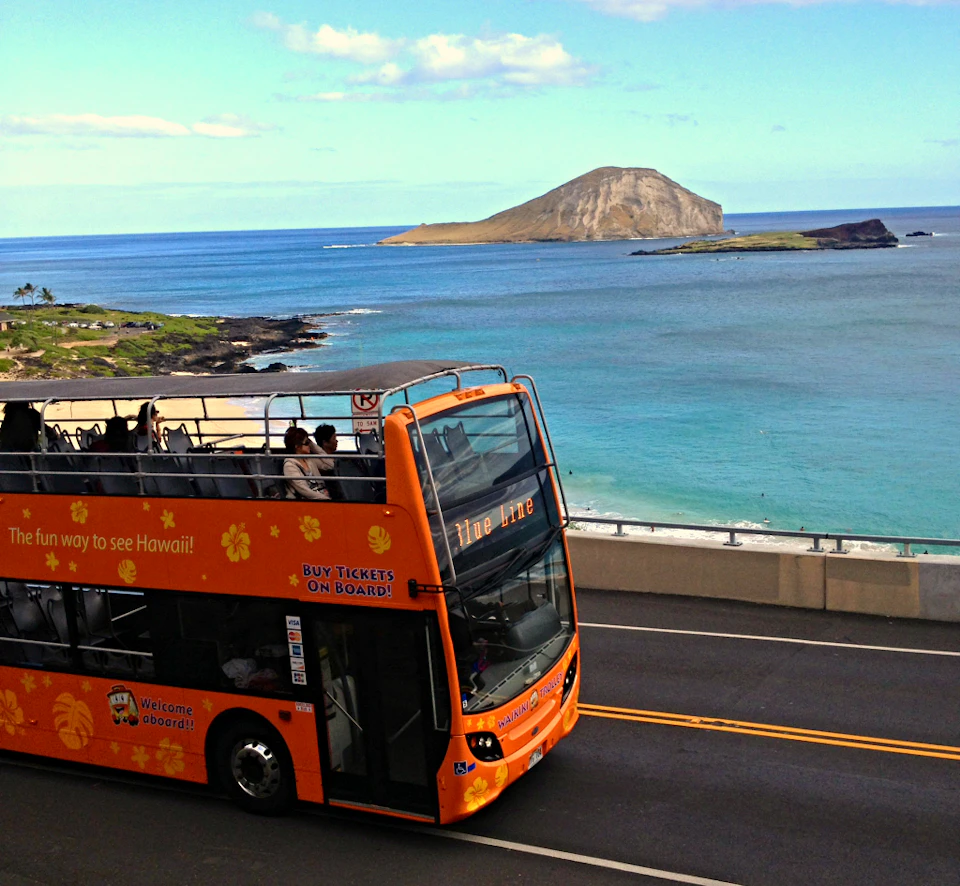 Open-top tour bus on a coastal road, blue ocean, small island, and partly cloudy sky in the background.