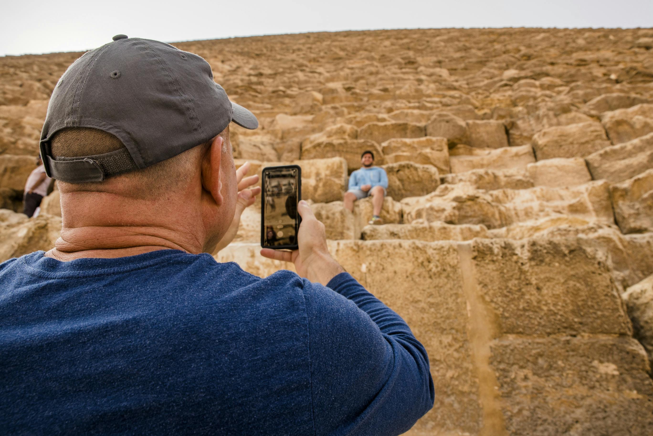 Person wearing a gray cap and blue shirt taking a photo with a smartphone of another person sitting on large stone steps.