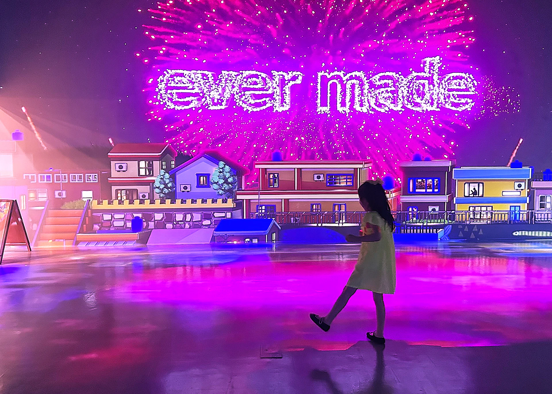 A child in a yellow dress walks in front of a colorful backdrop of houses and the words 'ever made' in fireworks.