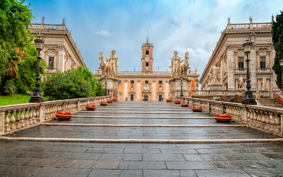 Wide stone staircase flanked by statues and flower pots leads to a historic building with a central clock tower under a blue sky.