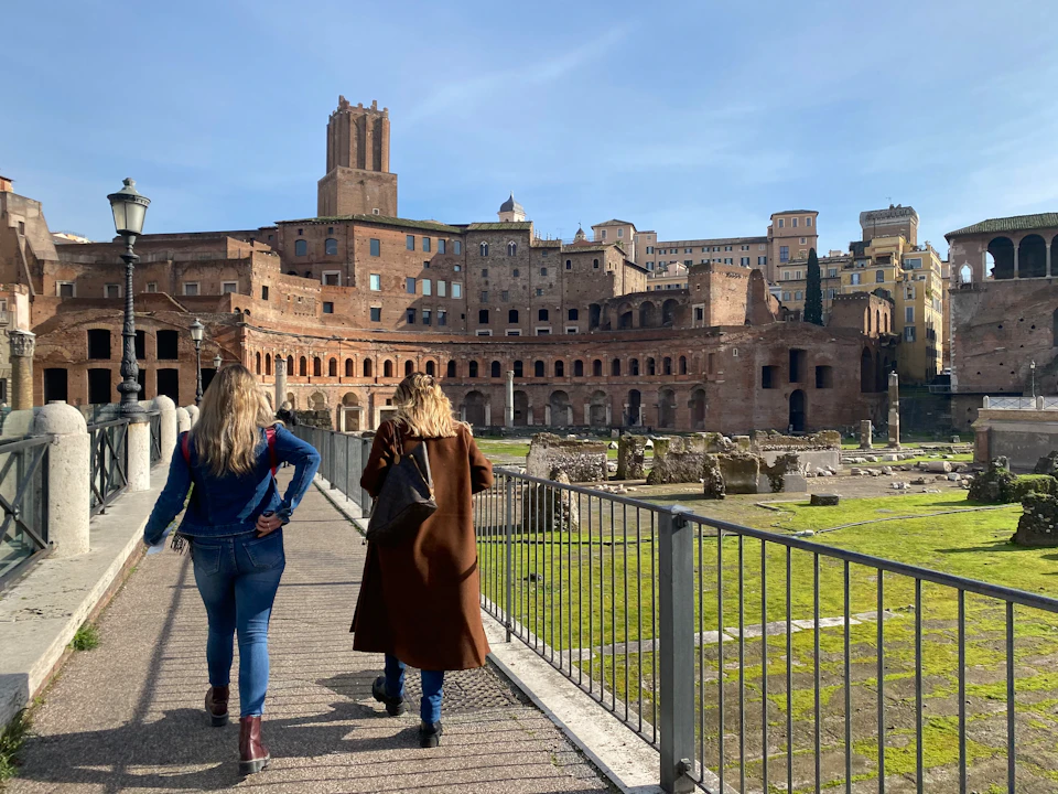 Two people walk toward ancient Roman ruins with red-brick structures, under a clear blue sky.