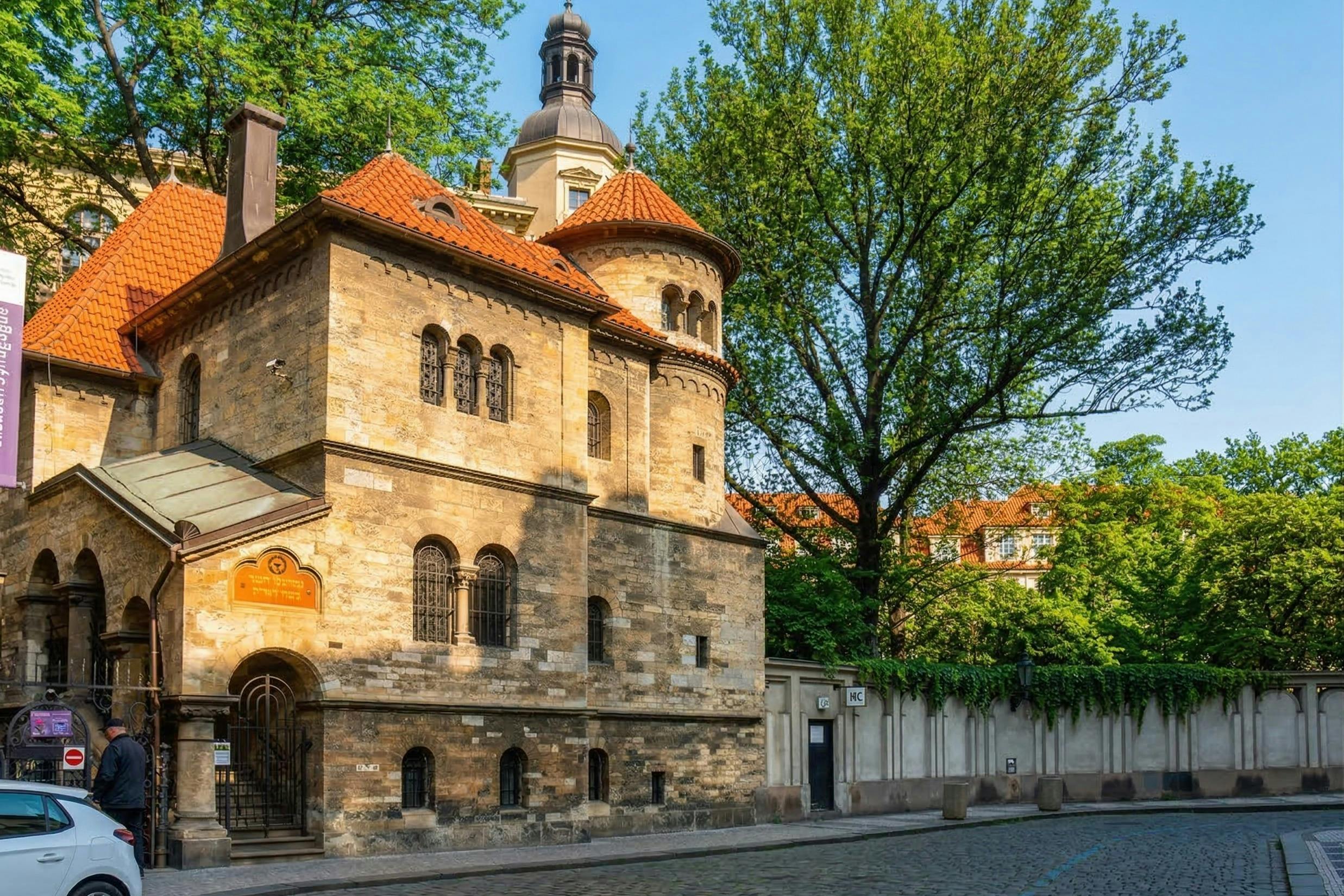Historická kamenná budova s klenutými okny a červeně obloženou střechou, obklopená stromy a dlážděnou ulicí.