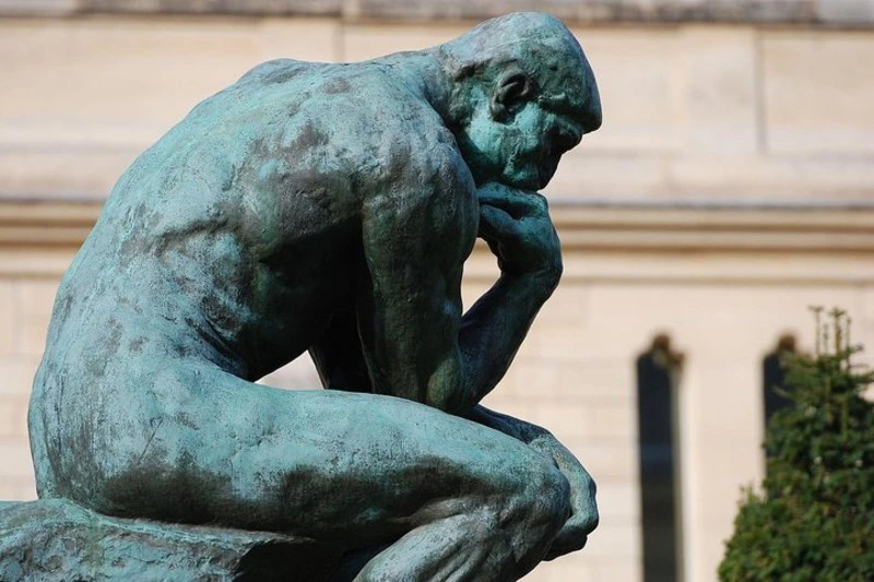 Bronze statue of a seated man, resting chin on his hand, in a thoughtful pose, with a blurred architectural backdrop.