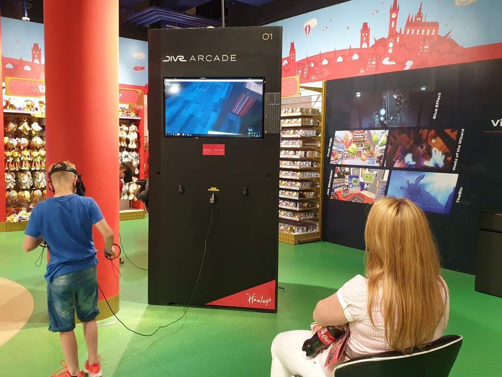 Ребенок использует VR-гарнитуру на аркадной станции Hamleys VR, в то время как женщина смотрит в красочном магазине игрушек.