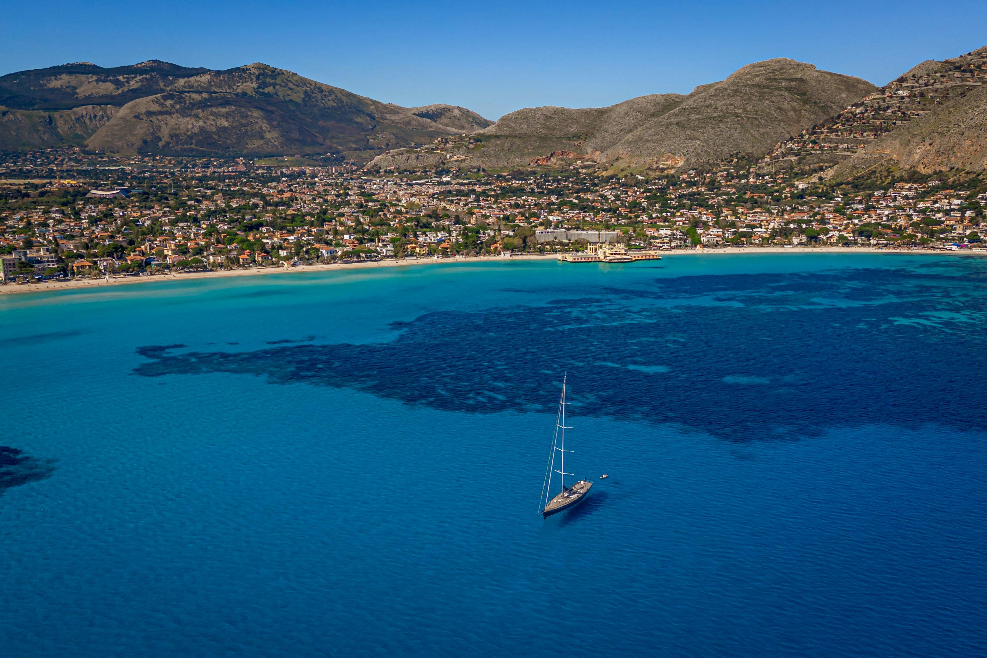 Vue aérienne d'une baie avec un voilier dans une eau bleue claire, avec une ville côtière et des montagnes en arrière-plan.