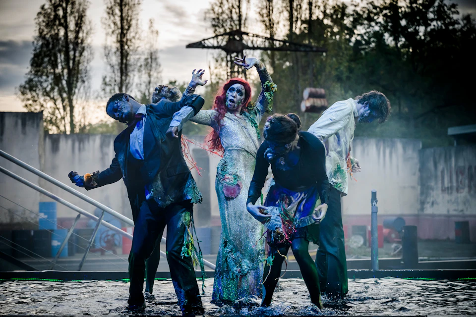 A group of performers in zombie costumes act dramatically on a foggy outdoor stage with trees in the background.