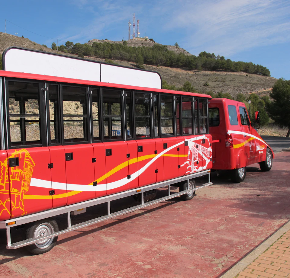 Red tourist tram with black windows parked on a paved area, with hills and trees in the background under a blue sky.