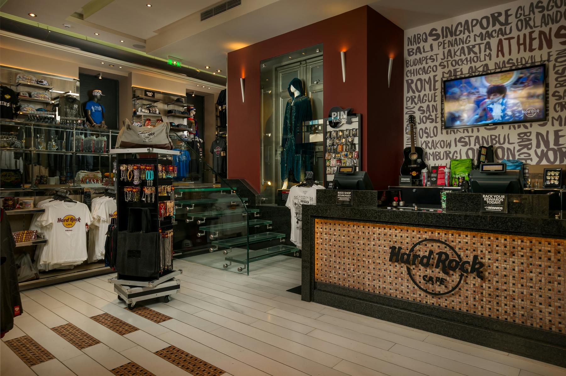 Interior of a Hard Rock Cafe shop with merchandise, including T-shirts, a display case with pins, glass stairs, and a counter with a guitar.