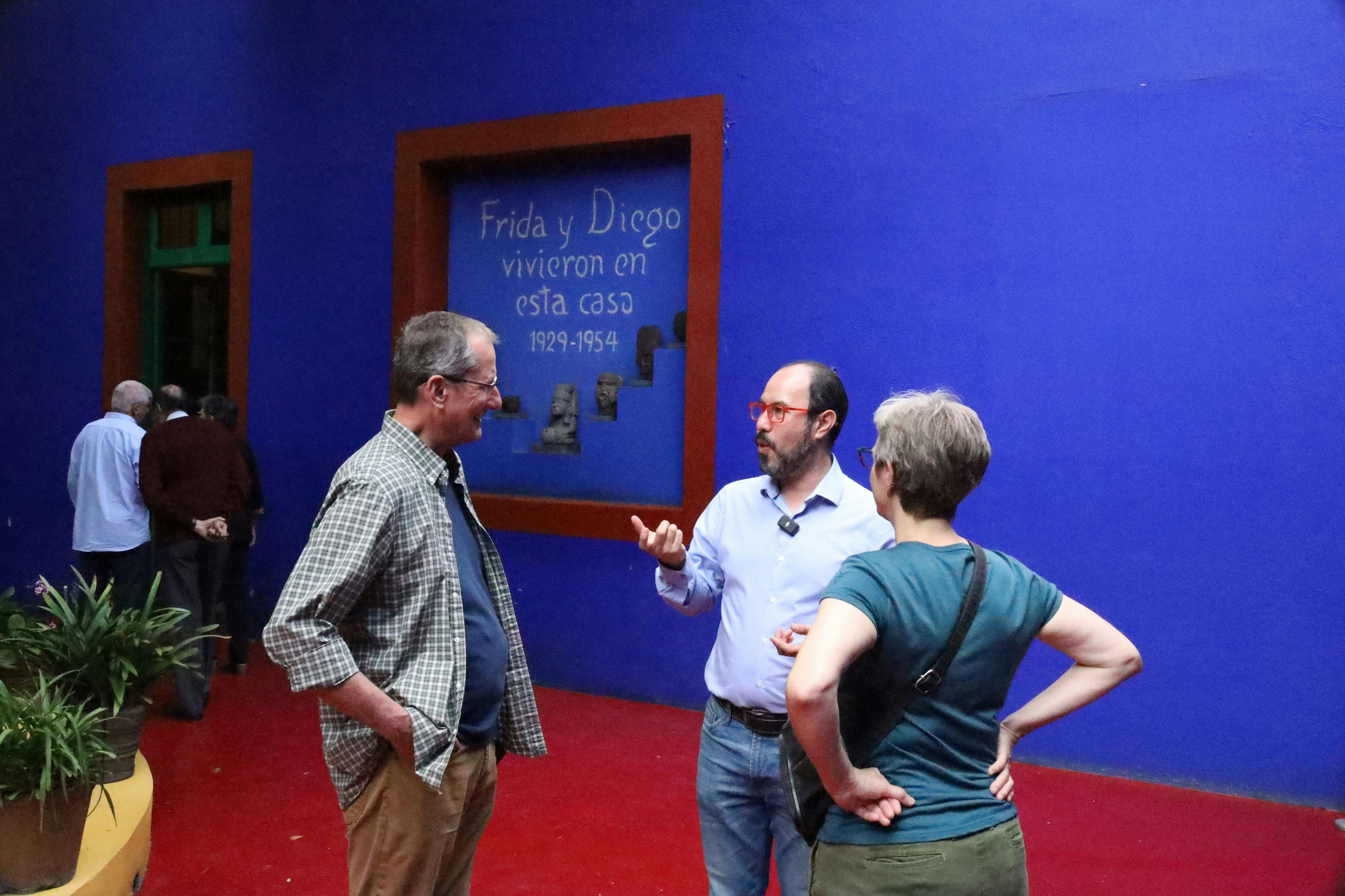 Drei Personen unterhalten sich vor einer blauen Wand mit einem Text über Frida und Diego, im Vordergrund ein roter Boden.