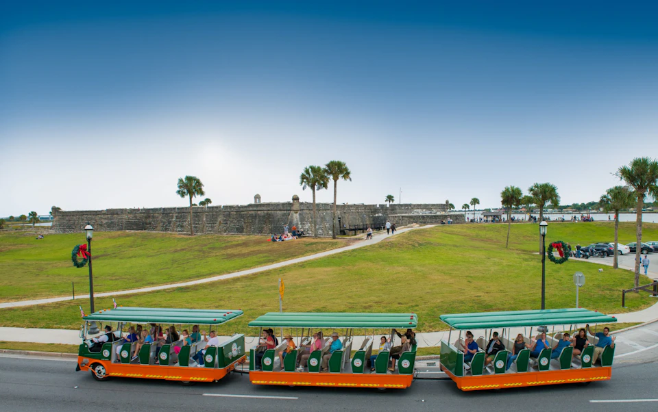 A tram with tourists passes by a historic stone fort with grassy lawns and palm trees under a clear blue sky.