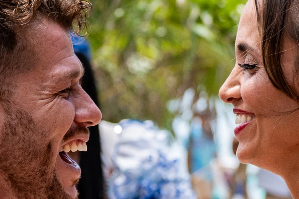 A man and a woman laugh and hold hands outdoors, with people and palm trees in the background.