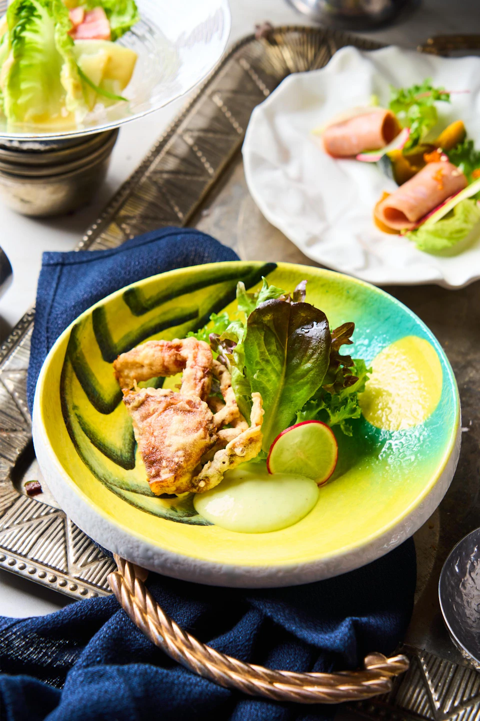 A colorful dish with fried soft-shell crab, leafy greens, radish slices, and a dollop of sauce on a vibrant ceramic plate.