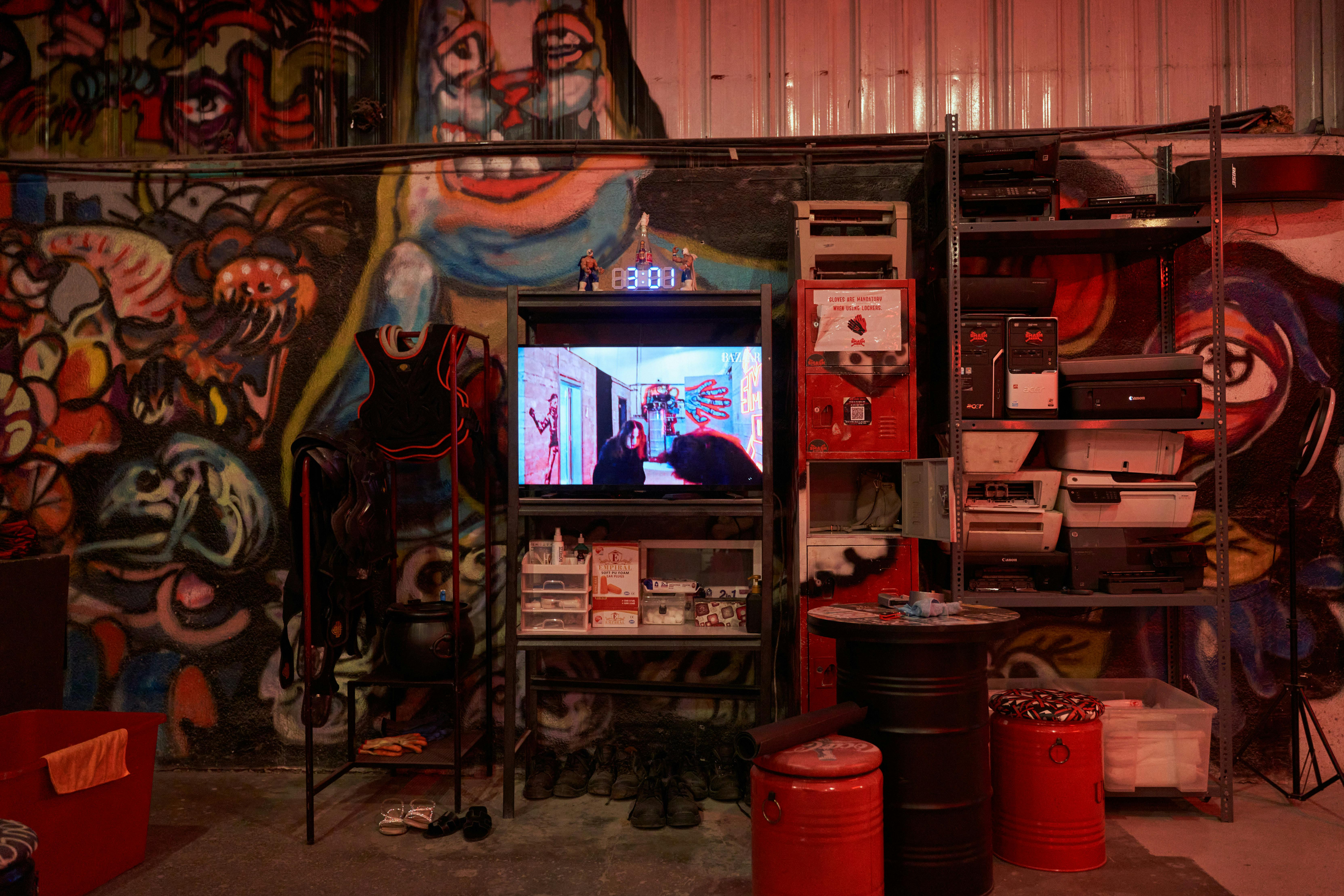 Δωμάτιο με ζωηρά γκράφιτι στον τοίχο, μεταλλικό εξοπλισμό συγκράτησης στο ράφι, τηλεόραση που προβάλλει μια σκηνή, κάδους αποθήκευσης και διάφορα αντικείμενα.